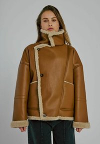 Vrouw in een tan kleurige leren jas met schapenvachtvoering, asymmetrische knoopsluiting en hoge kraag, staande voor een effen grijze achtergrond.
