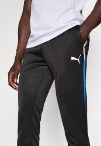 Pantaloni de sport negri cu accente albastre pe lateral și un logo Puma alb. Realizați dintr-un material fin și ușor, cu un croi lejer și buzunare.