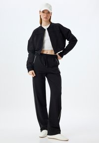 Chaqueta negra recortada con dos bolsillos frontales, combinada con pantalones deportivos negros que tienen franjas laterales blancas y una cintura elástica. Zapatillas blancas y gorra.