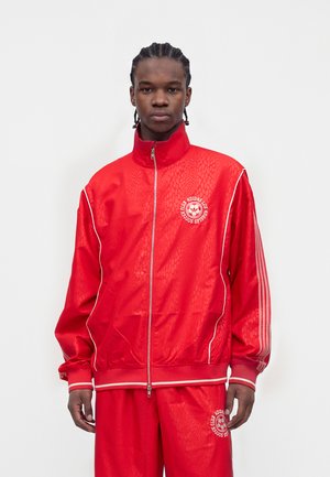 Jeune homme aux cheveux tressés portant un survêtement rouge du club de football Adidas avec des liserés blancs et un logo, debout devant un fond uni.