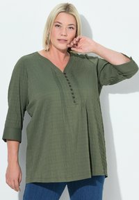 Olivgrüne Bluse mit V-Ausschnitt, dreiviertellangen Ärmeln und strukturiertem Finish. Mit einer Knopfleiste vorne.