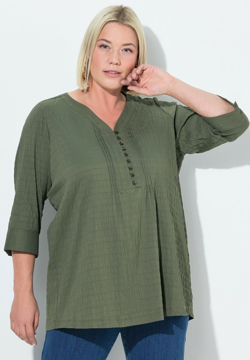 Olivgrüne Bluse mit V-Ausschnitt, dreiviertellangen Ärmeln und strukturiertem Finish. Mit einer Knopfleiste vorne.