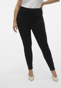 Jeans negros ajustados con una textura suave, cintura alta, bolsillos frontales y cierre con botón, combinados con tacones puntiagudos de color claro.