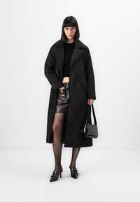 Woman in black long coat, black cropped top, dark brown leather mini skirt, black sheer tights, black heels, holding black handbag.