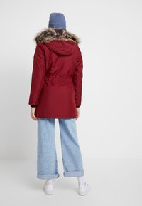 Röd parkajacka med fuskpäls på huven, figursydd midja och långa ärmar, i kombination med ljusblå vida jeans och vita sneakers.