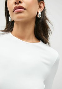 Vrouw die grote, glanzende, druppelvormige zilveren oorbellen draagt en een witte ronde hals top, zichtbaar van de kin tot de bovenkant van de borst.