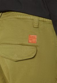 Maslinaste hlače od glatkog materijala, s stražnjim džepom s ušivenim poklopcem i kvadratnom narančastom oznakom "Alpha Industries."