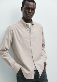 Camicia beige con bottoni realizzata in tessuto morbido, con colletto classico e maniche lunghe, abbinata a jeans grigio scuro. Design e texture semplici.