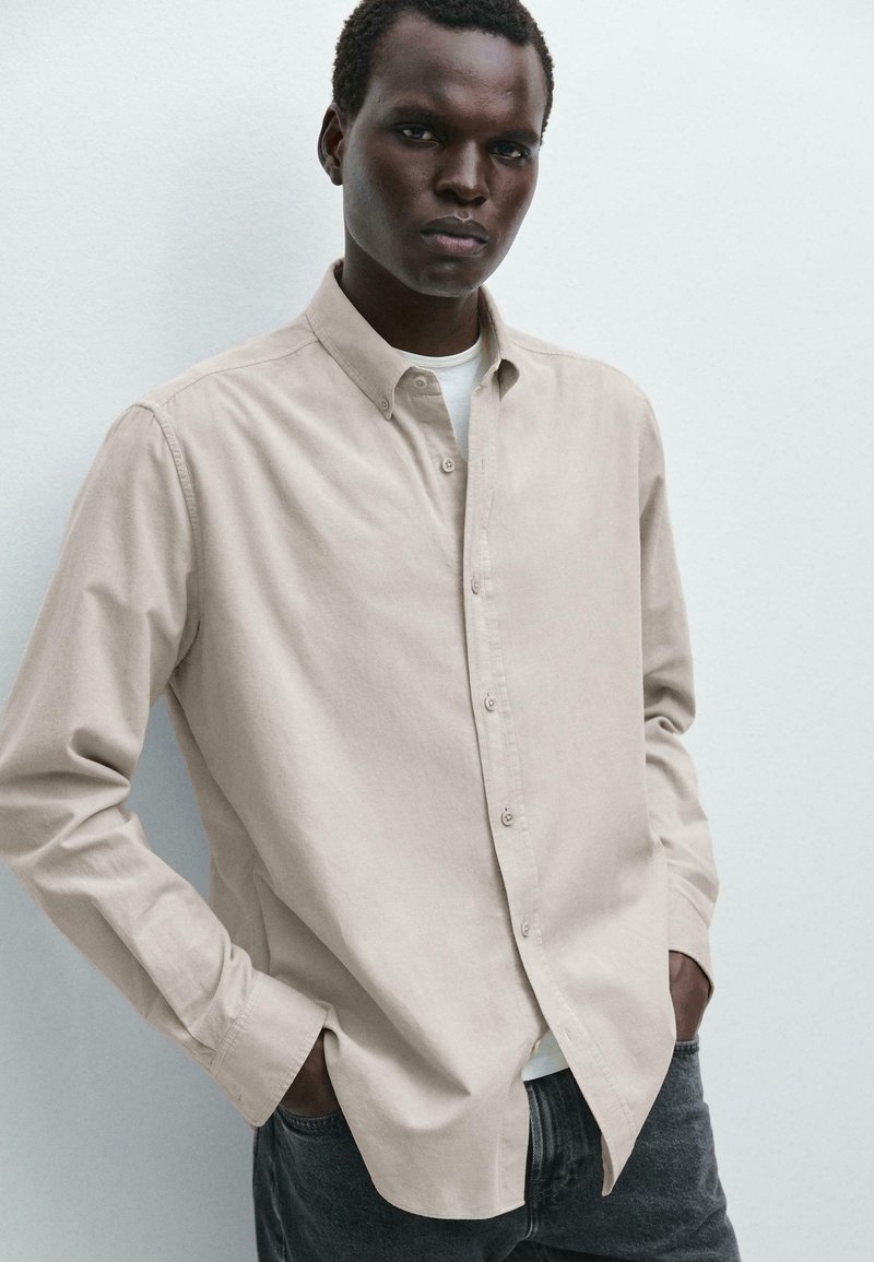 Camicia beige con bottoni realizzata in tessuto morbido, con colletto classico e maniche lunghe, abbinata a jeans grigio scuro. Design e texture semplici.