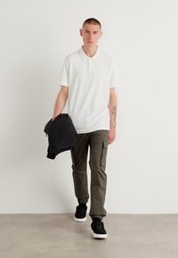 Hvit polo shirt, oliven cargo bukser og svarte joggesko. Modell som holder en svart jakke. Nøytral bakgrunn, minimalistisk stil.