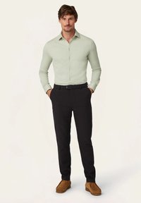 Lichtgroen, lange mouwen blouse met knopen, gecombineerd met zwarte broek. Model draagt bruine instappers. Effen achtergrond.