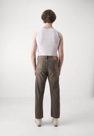 Personne debout de dos, portant un débardeur blanc, un jean marron et des baskets blanches montantes sur un fond uni.