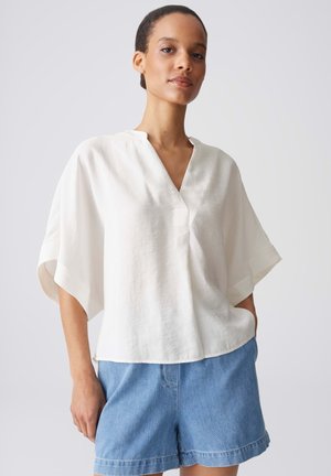Junge Frau trägt eine lockere weiße V-Ausschnitt-Bluse mit weiten Ärmeln und helle blaue Jeansshorts, steht mit einer Hand in der Tasche.