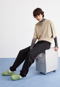 Khaki oversized tričko, černé tapered kalhoty, zelené slip-on dřeváky. Sedí na stříbrném kovovém kontejneru s kolečky. Texturovaný povrch, lehký styl.