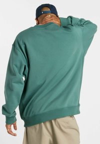 Sweat-shirt vert avec une coupe décontractée, des poignets et un ourlet côtelés, et un tissu texturé. Le dos présente un design simple sans logos visibles.