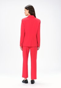 Completo su misura rosso, con giacca a un bottone e rever a scomparsa e pantaloni dritti abbinati. Tessuto liscio e hardware minimal.