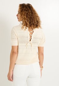 Top de ganchillo crema con mangas cortas, espalda descubierta que presenta un detalle de cordones y un patrón texturizado. Combinado con pantalones blancos de cintura alta.