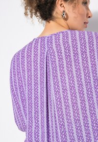 Lilla bluse med et subtilt stjernemønster og lodrette striber. Inkluderer en rygplyt for ekstra designdetalje. Glat stofstruktur.