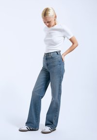 Los jeans de mezclilla anchos de color azul claro presentan una cintura alta, diseño de cinco bolsillos y una textura desgastada. Combinados con una camiseta blanca ajustada y zapatillas.