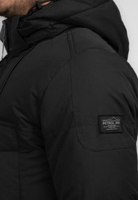 Giacca a vento nera con cappuccio, finitura testurizzata e una distintiva patch con logo sulla manica sinistra. Cuciture rinforzate visibili sulle giunture.