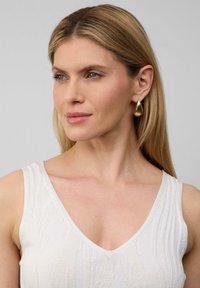 Złote kolczyki w kształcie kropli z polerowanym wykończeniem, noszone z białą teksturowaną odzieżą z dekoltem w serek. Gładkie włosy opadają na ramię.