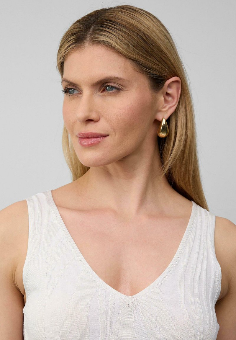 Złote kolczyki w kształcie kropli z polerowanym wykończeniem, noszone z białą teksturowaną odzieżą z dekoltem w serek. Gładkie włosy opadają na ramię.