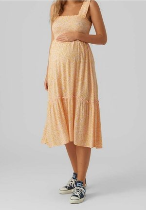 Schwangere Frau in einem ärmellosen, pfirsichfarbenen Midikleid mit Blumenmuster und schwarzen High-Top-Sneakers, die sanft ihren Bauch hält.