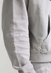 Light gray sweatshirt sleeve with subtle embossed floral and cross design, ribbed cuff, and pocket detail on the side.