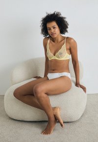 Lacy yellow bra with thin straps, paired with white panties featuring a lace trim. Model seated on a textured beige chair.