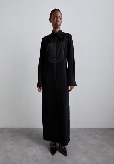 Robe noire longue à boutons en tissu lisse avec un col arrondi et des poignets évasés, associée à des chaussures noires à bout pointu.