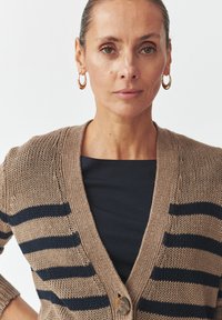 Tan knitted cardigan with navy stripes, v-neck design, and a front button closure. Worn over a black top, featuring ribbed cuffs.