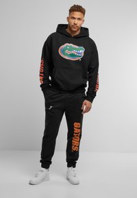 Sudadera negra con un logotipo de caimán colorido, texto naranja "GATORS" en las mangas; combinada con pantalones de chándal negros que presentan la marca "GATORS" en naranja.