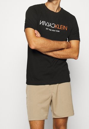 Man met een zwarte Calvin Klein T-shirt en beige korte broek, met gekruiste armen tegen een effen achtergrond.
