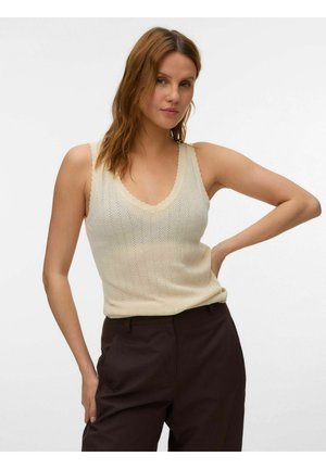 Femme aux cheveux châtain clair portant un haut en tricot crème sans manches et un pantalon marron foncé, posant avec une main sur la hanche devant un fond uni.