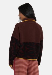 Brązowa kurtka polarowa z czarnym abstrakcyjnym wzorem, żółty akcent przy kołnierzu oraz luźny krój, z rękawami raglanowymi i teksturowaną powierzchnią.