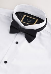 Bílá košile s černým motýlkem. Má špičatý límec, lesklá zapínání na knoflíky a texturovanou černou podšívku s bílými puntíky.