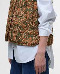 Gevoerde gilet in olijfgroen met ingewikkelde koperen bloemmotieven, uitgerust met een zak en Barbour-logo, gedragen over een wit shirt.