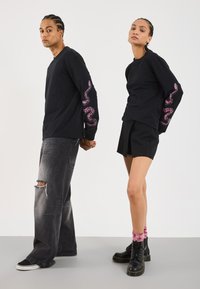Des chemises noires à manches longues présentent des graphiques de serpents roses sur les manches. À porter avec un jean noir déchiré et une jupe noire, complétés par des bottes de combat.