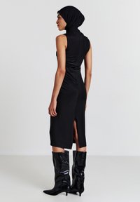 Abito nero senza maniche con collo alto, silhouette elegante e spacco posteriore, abbinato a stivali neri lucidi sopra il ginocchio con punta affusolata.