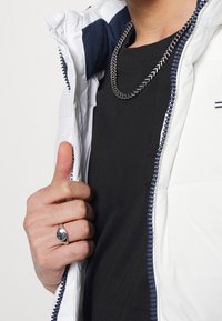 Vit puffjacka med marinblå vaddering, svart T-shirt under, silverkedja runt halsen och en reflekterande silvring på en hand.