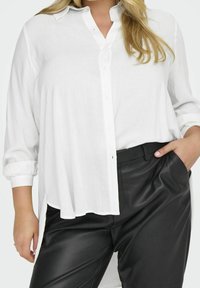 Camicia bianca con bottoni, maniche arrotolate, in tessuto leggero e con spacchi laterali, abbinata a pantaloni in pelle nera. Texture liscia con vestibilità classica.