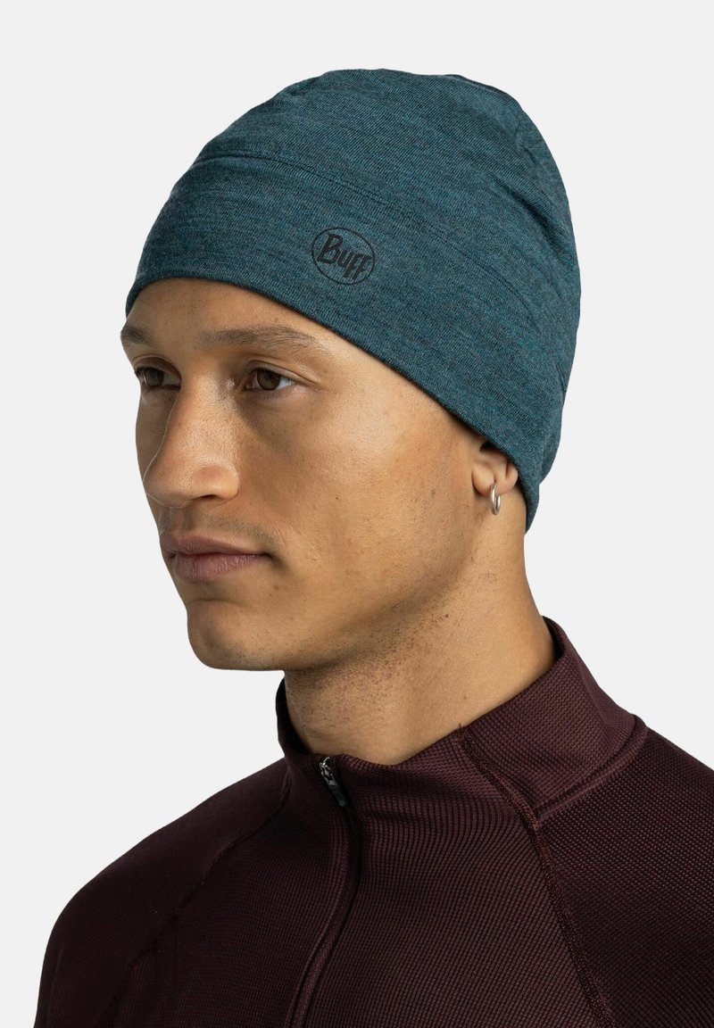 Young man wearing a teal Buff beanie and maroon zip-up textured jacket, facing left with a neutral expression and small hoop earring.