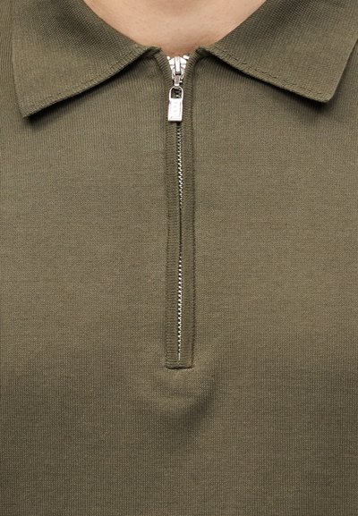 Πλεκτό πόλο σε ελιά πράσινο χρώμα με μερικώς φερμουάρ ασημί φερμουάρ και διπλωμένο γιακά στον λαιμό και το πάνω μέρος του στήθους ενός ατόμου.