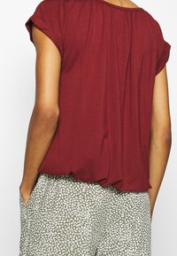 Burgundy kortärmad topp med lös passform och rynkad halsringning, matchad med ljus olivgröna byxor med ett vitt bladmönster.