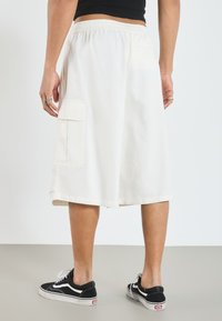 White cotton shorts with an elastic waistband and two side pockets, styled just above the knee. Paired with black sneakers.
