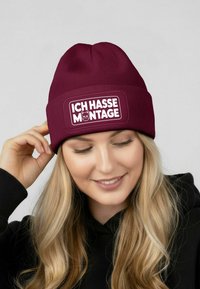Maroon beanie with ribbed texture, featuring a rectangular white patch that reads "ICH HASSE MONTAGE" with a cat illustration.