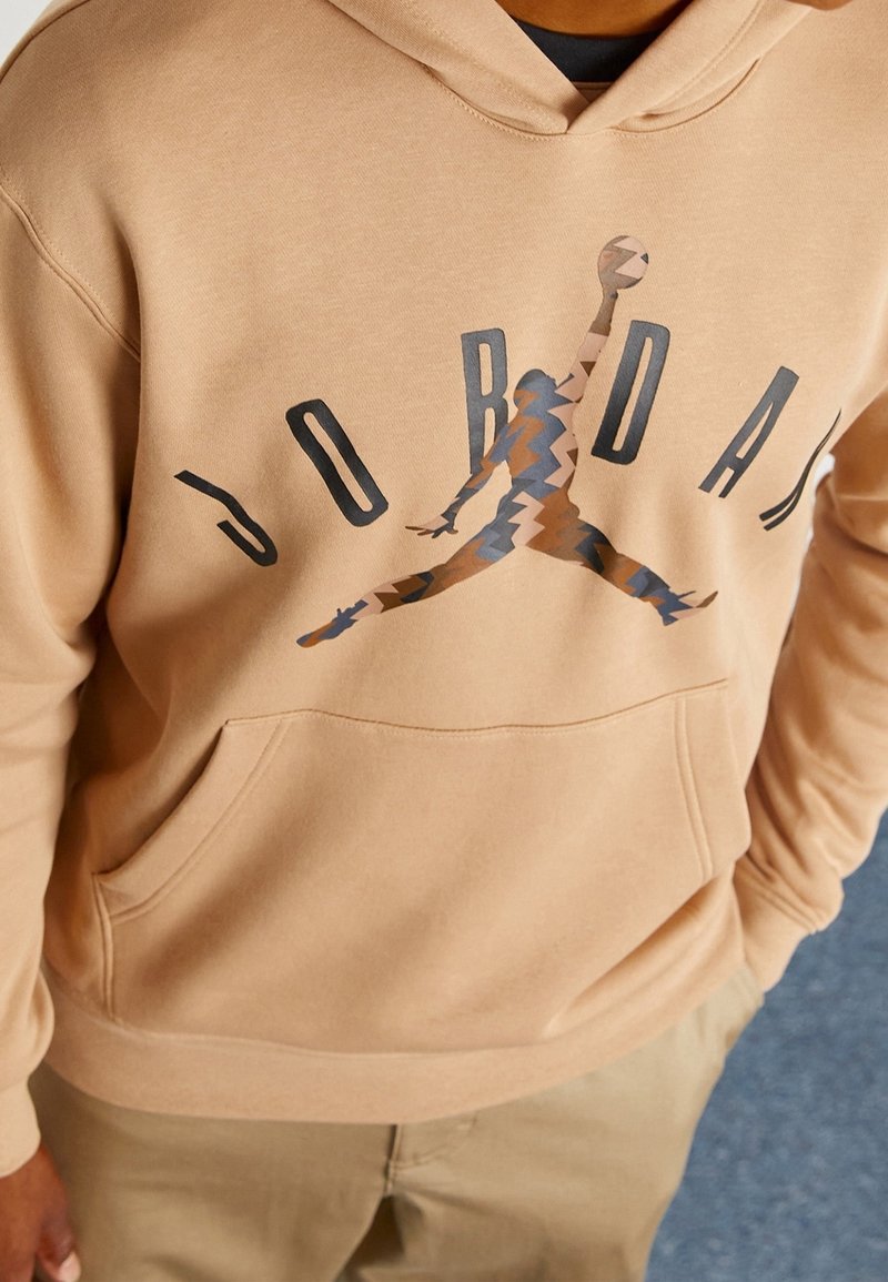 Personne portant un sweat à capuche beige avec un logo Jordan stylisé comprenant la silhouette d’un joueur de basketball et de grandes lettres imprimées sur la poitrine.