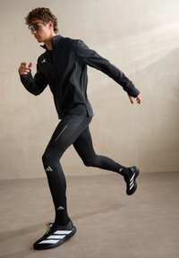 Junge Person in schwarzer Sportbekleidung und Sonnenbrille läuft drinnen auf einem beigen Boden und vor beiger Wand.
