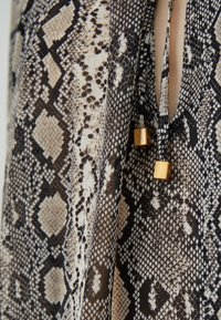 Slangenhuidpatroon stof in beige en zwart. Heeft dunne bandjes met gouden vierkante accenten. Gelaagde oppervlakte met een mix van schubben en vormen.