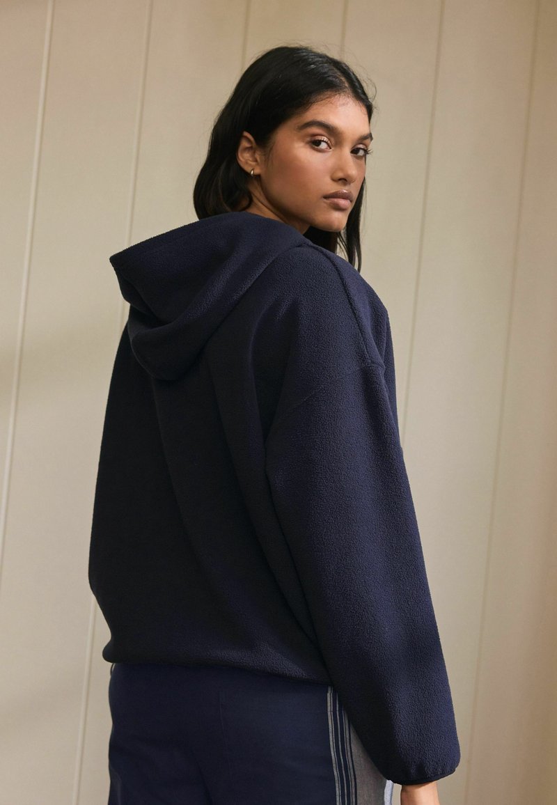 Mörkblå hoodie i mjukt, texturerat fleecematerial med sänkt axelparti. Har en avslappnad passform och ribbade muddarna.